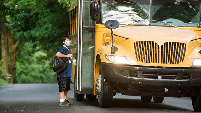 孩子带着棕色的头发和面具，在敞开的校车门前背着书包