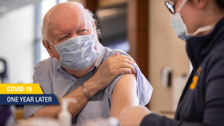 一名白发老人戴着蓝色口罩，戴着蓝色衬衫，从工作人员那里接种疫苗