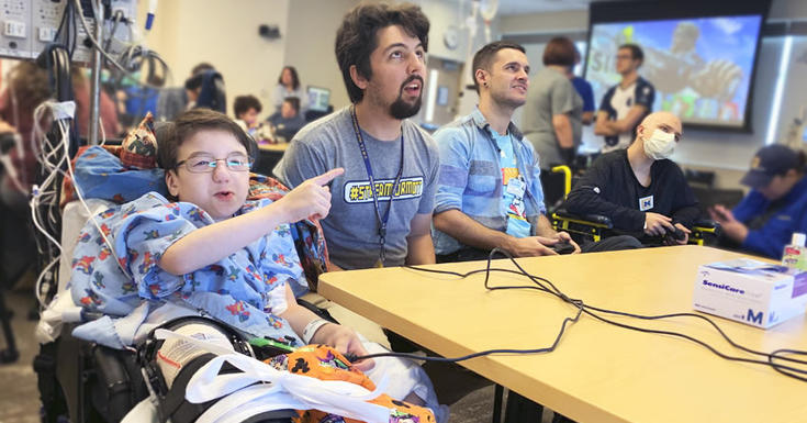 指向从轮椅的孩子到有视频游戏治疗师的录影屏幕玩与遥控在他旁边的遥控在桌子上满是人