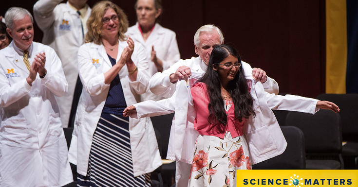 身着白大褂的医生们站在舞台上对着人群给女学生穿白大褂