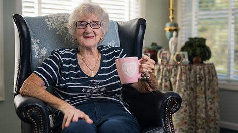 奶奶面带微笑，手里拿着粉红色的杯子坐在椅子上
