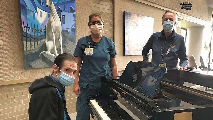 一名戴口罩的男子坐在钢琴旁，戴口罩的护士站在他身边，一名戴口罩的男子站在钢琴对面