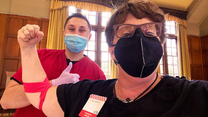masked woman with glasses smilling with red gauze on arm and nurse standing behind masked giving thumbs up